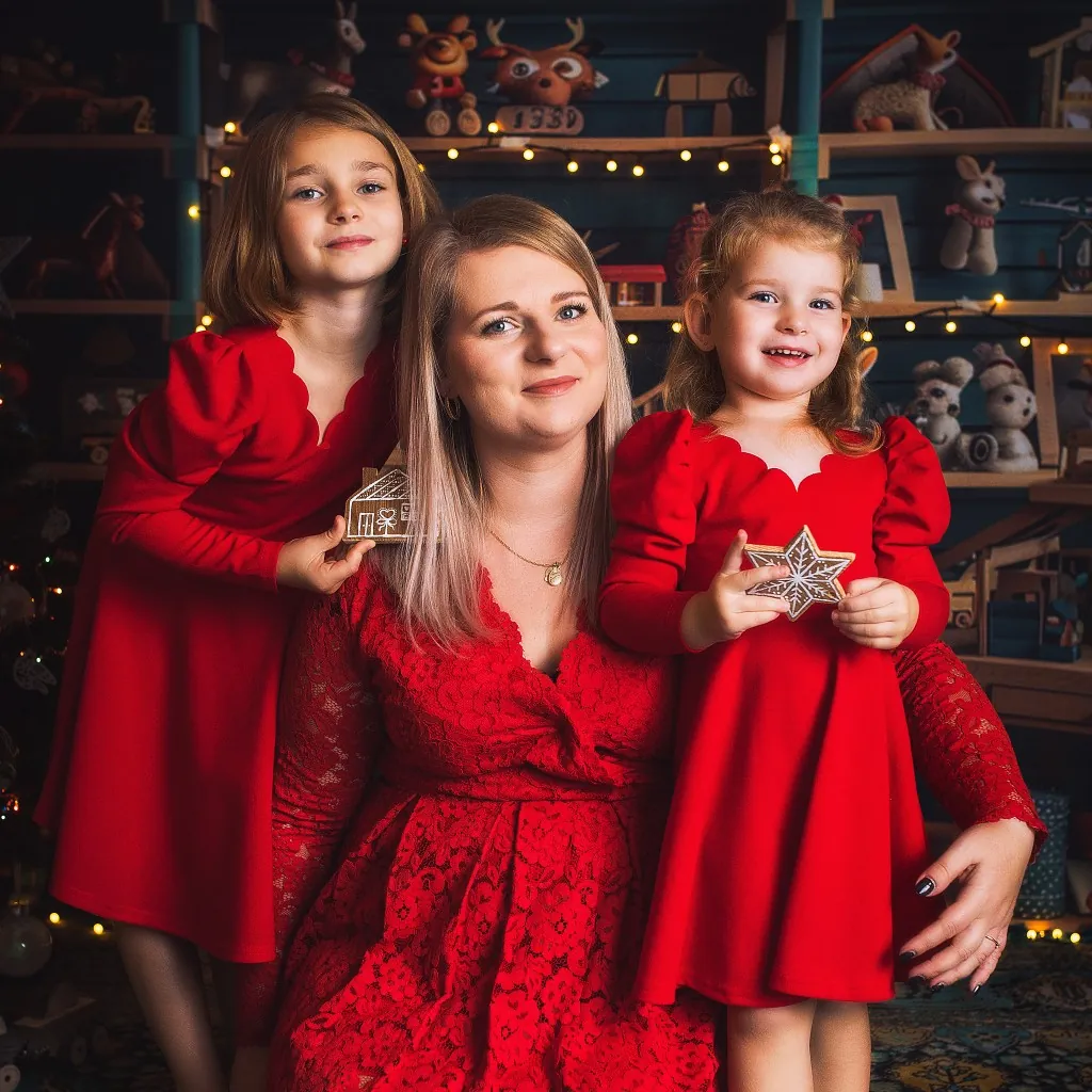 Mama w czerwonej koronkowej sukience z dwiema córkami, które trzymają piernikowy domek i gwiazdkę.
